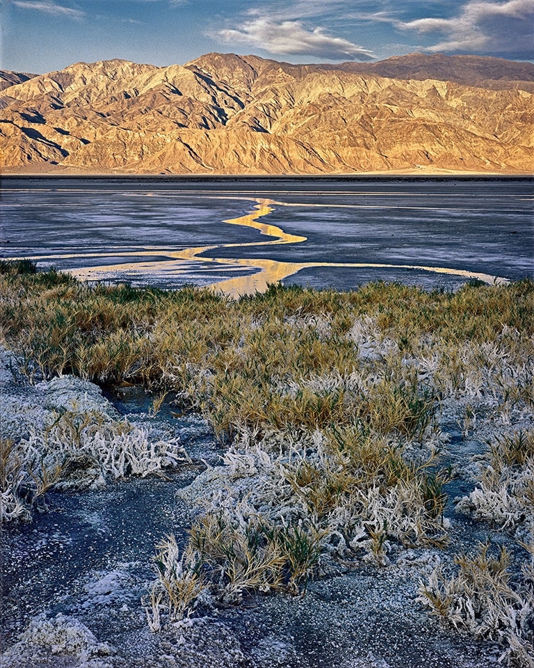 The just risen sun reddens the distant mountain which is normally almost colorless.  Salt clings to the salt grass.  In between the unlikely water standing in this dry place reflects the mountains on a palette of blue mud.  Death Valley National Park, CA.